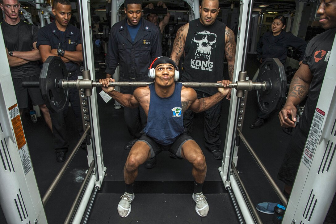 A sailor squats while holding a barbell on his shoulders as others observe.