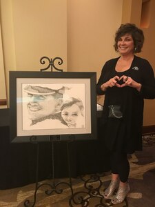Janine Sijan-Rozina forms a heart with her hands while standing next to a portrait of her and her brother, Capt.Lance Sijan. The portrait came from a screen capture of 8 mm footage of Sijan's Air Force Academy graduation. The artist, Charles Ingram, used the words “thank you” like brush strokes to create the image.