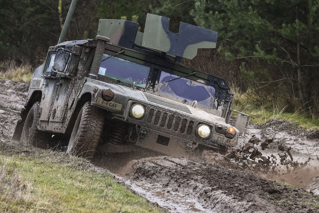 A camouflaged Humvee tilts and splashes up mud while driving on dirt roadway.