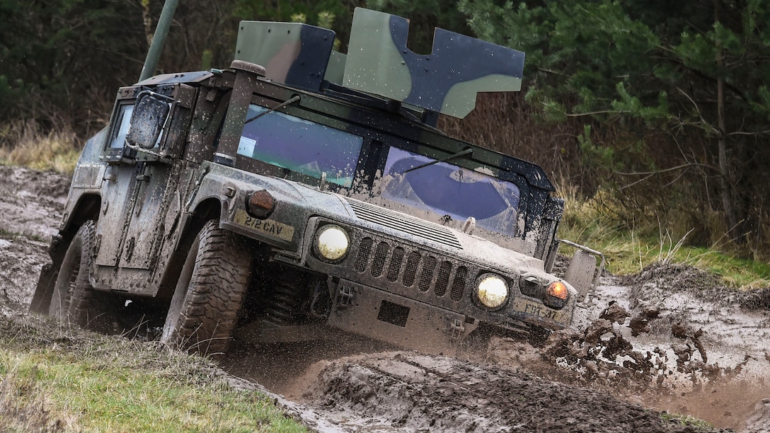 A camouflaged Humvee tilts and splashes up mud while driving on dirt roadway.