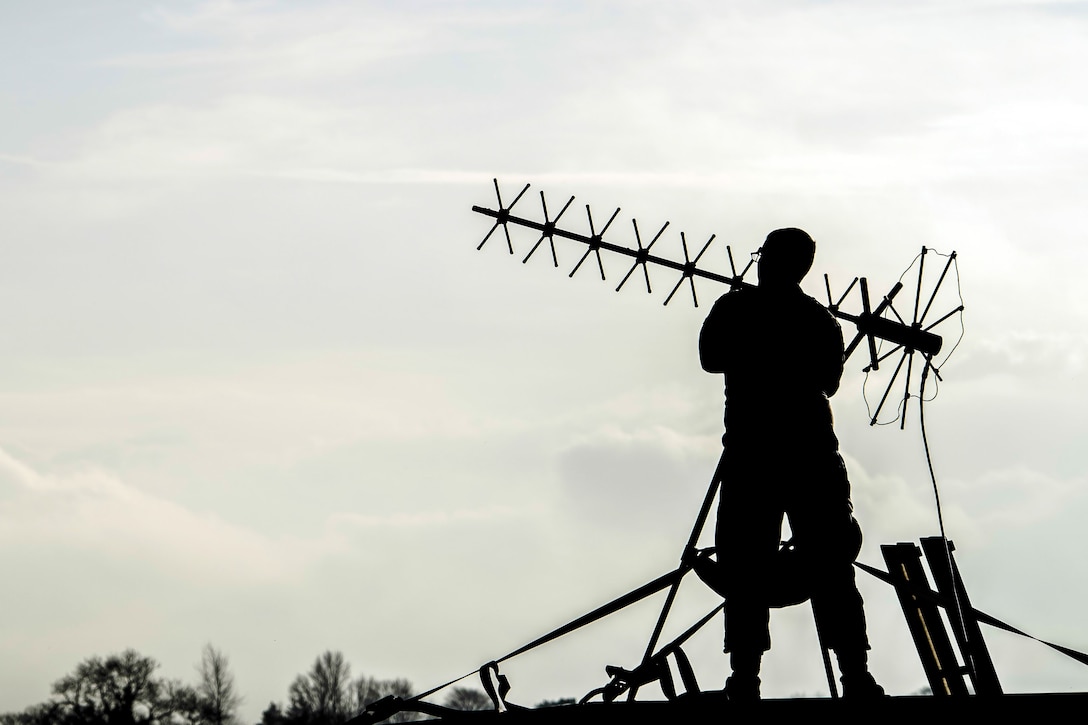 An airman, shown in silhouette, stands and adjusts an antenna.