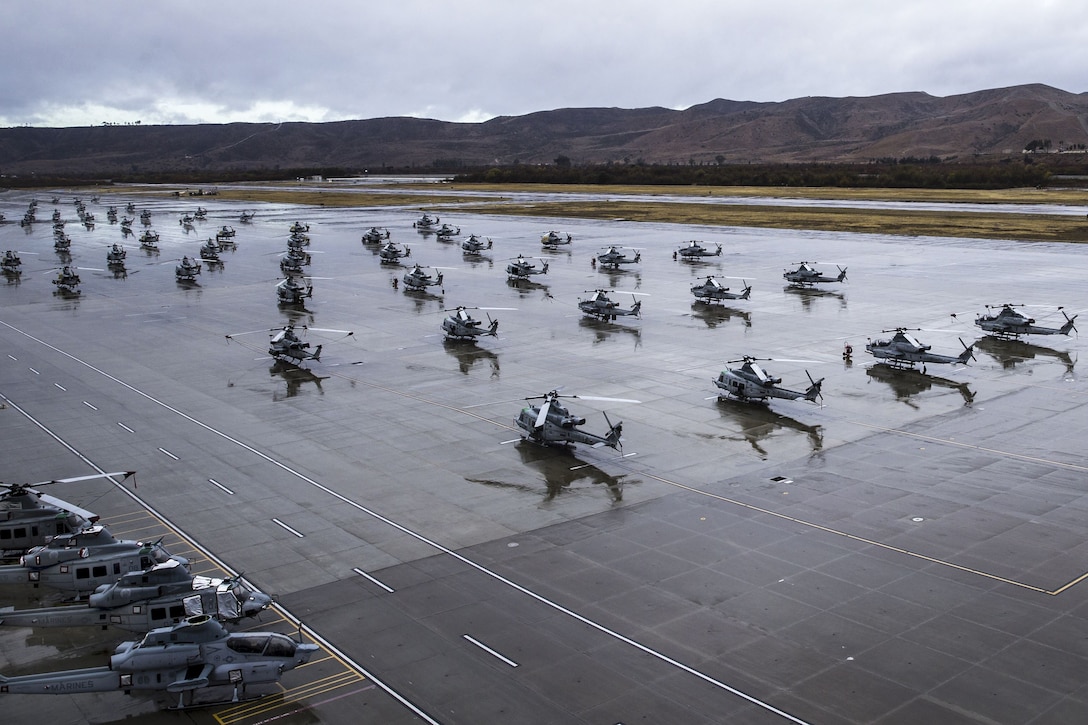 Rows of helicopters sit on a wet flightline, their reflections visible in the puddles on its surface.