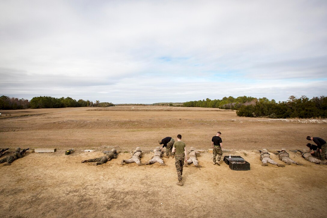 Marine recruits lay on the ground at a firing range with Marine instructors walking nearby.