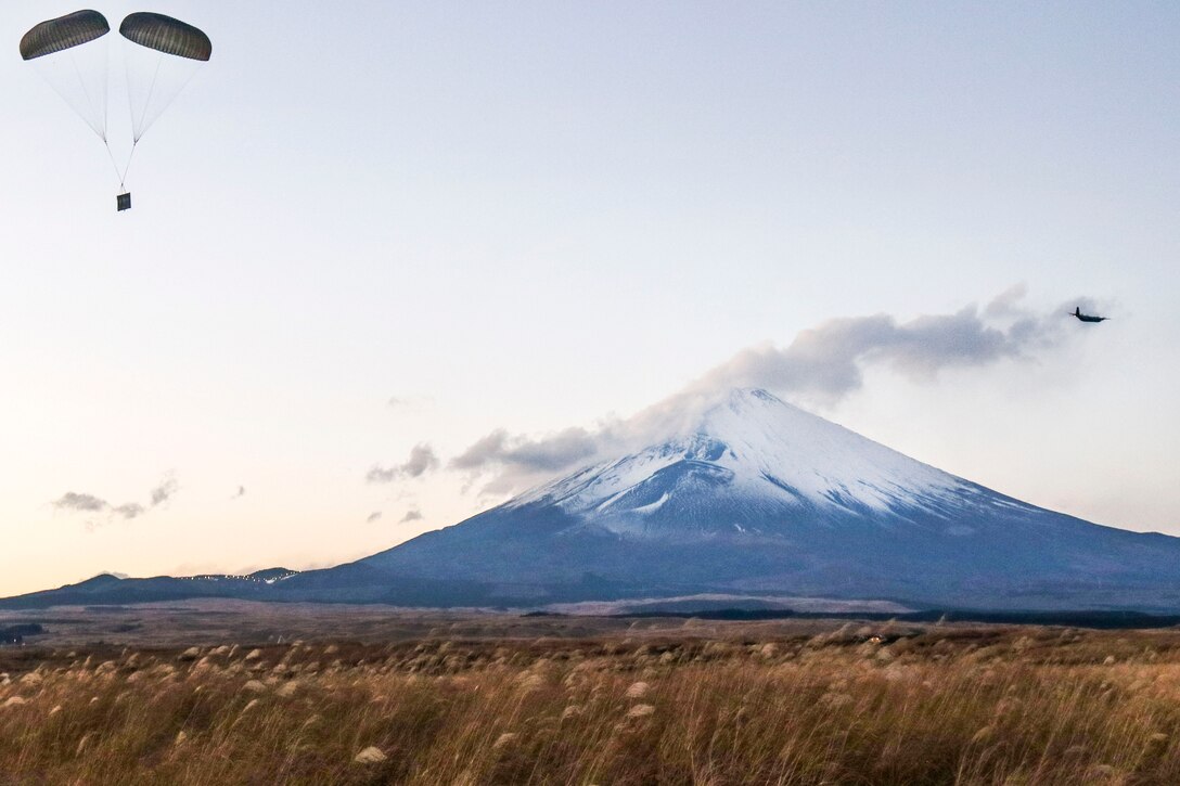 An aircraft flies by a mountain as a bundle drops from a parachute.