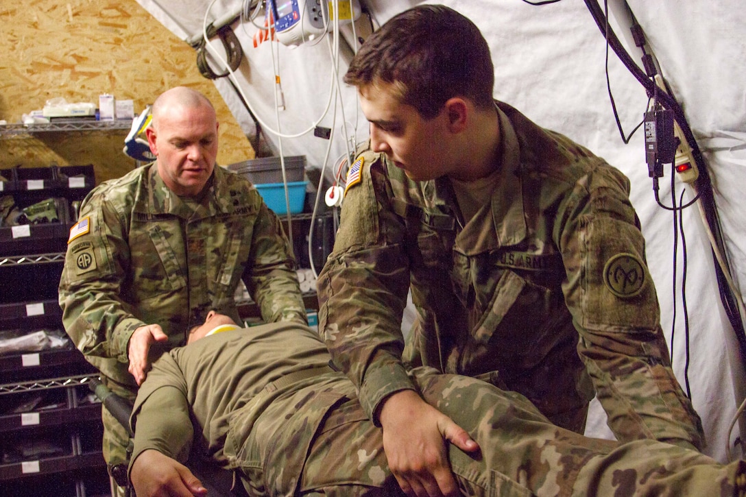Two soldiers lean over a soldier laying on a table.