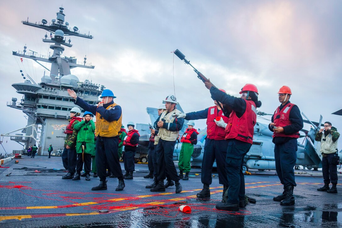 A sailor on a flight deck fires a shot line as others stand by.