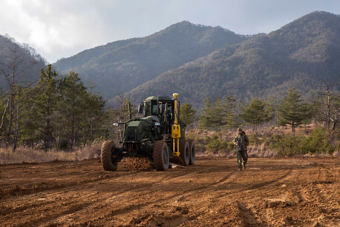 A Marine provides security and guides a heavy equipment operator leveling out terrain.