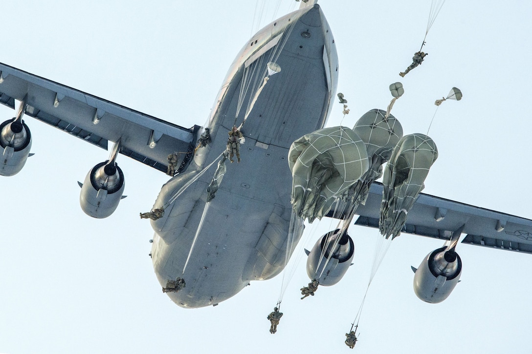 Soldiers with not-yet-open parachutes float around an aircraft in flight.