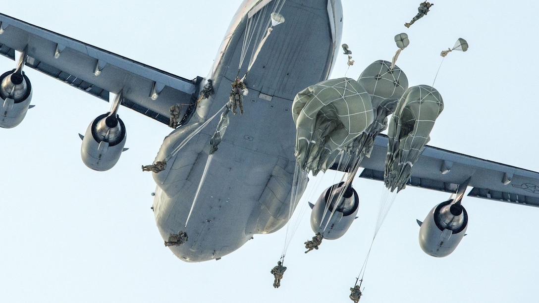 Soldiers with not-yet-open parachutes float around an aircraft in flight.