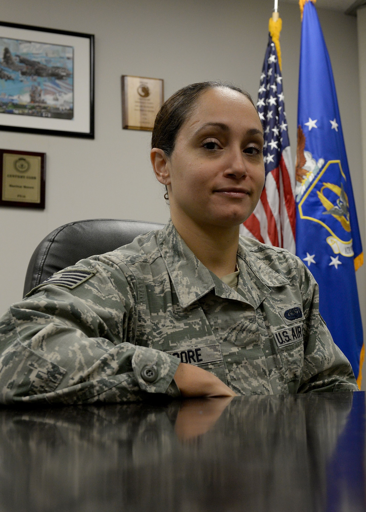 A woman in the Air Force Battle Uniform sits at a desk and smiles at the camera with an American Flag and the Reserve flag behind her.