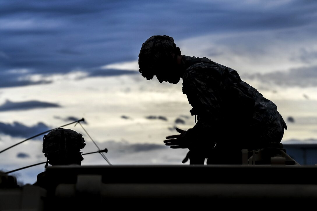 A soldier, shown in silhouette, leans over and motions while working on a weapon system with another soldier.