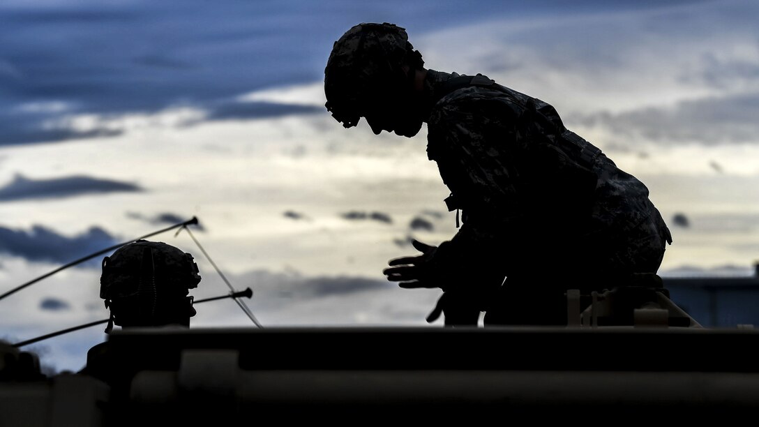 A soldier, shown in silhouette, leans over and motions while working on a weapon system with another soldier.
