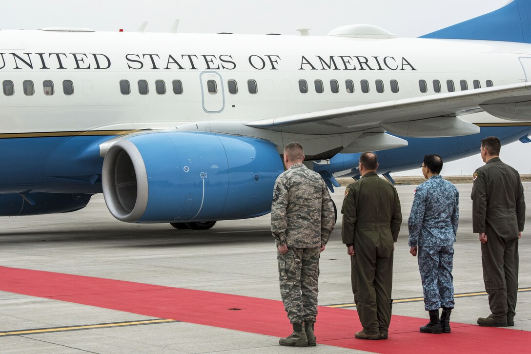 Four service members stand in a row by a red carpet and an aircraft on a flightline.