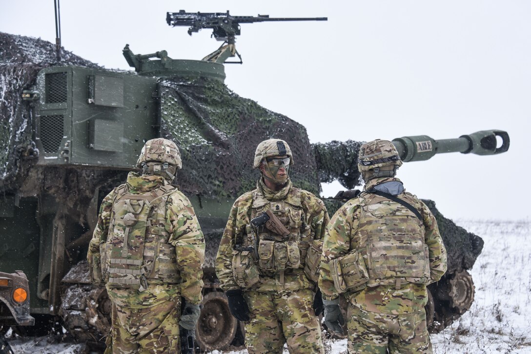 Three soldiers talking in front of a tank in a snow-covered field.