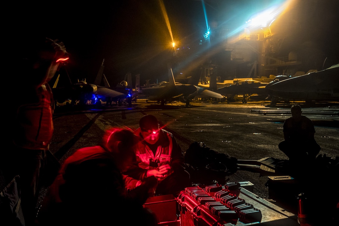 Sailors, illuminated by red light, work while sitting a ship's flight deck in darkness.