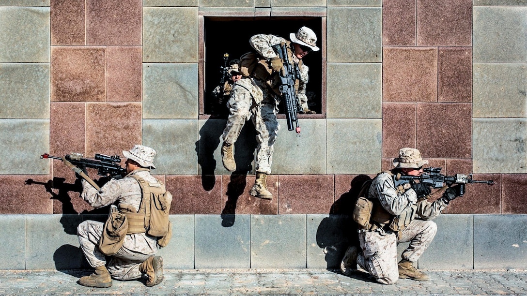 A Marine jumps through a building opening as two Marines kneel and point weapons outside it.