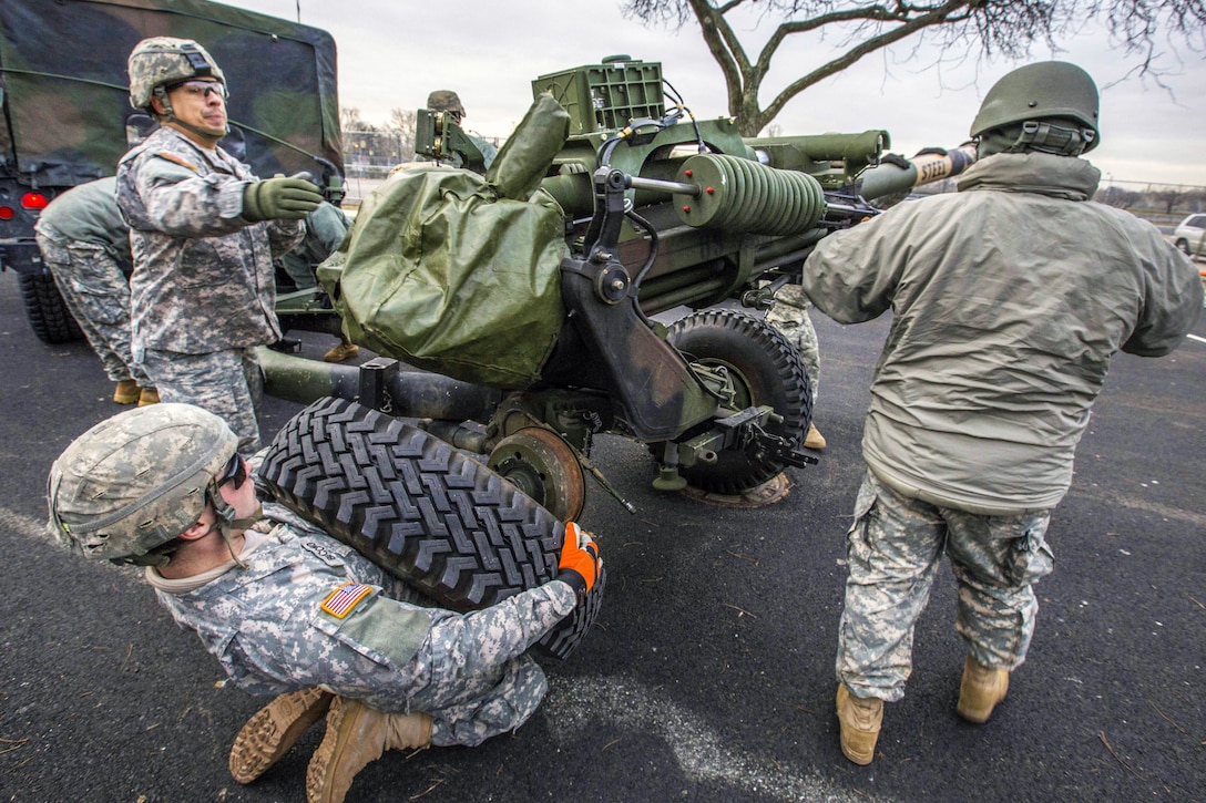 A soldier kneels and holds up a tire by a howitzer, as two others work near the weapon.