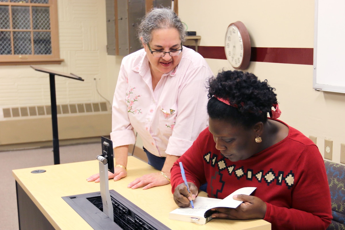 A woman watches as another woman signs a book at a desk.