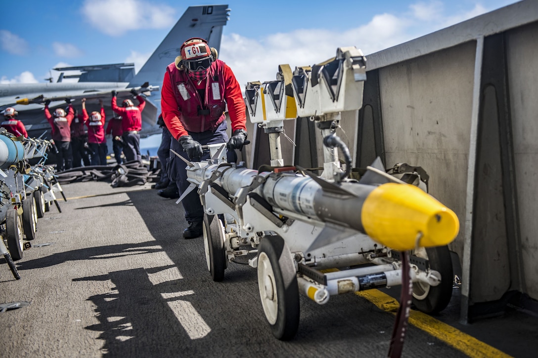 A sailor in a red uniform pushes a yellow-tipped missile on a wheeled cart on a ship's flight deck.