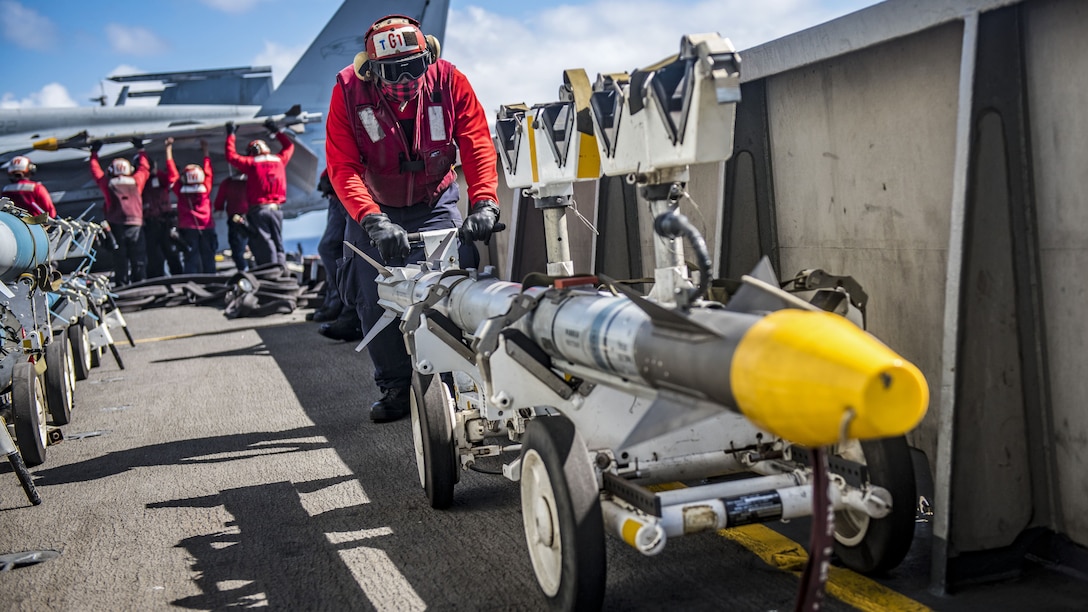 A sailor in a red uniform pushes a yellow-tipped missile on a wheeled cart on a ship's flight deck.