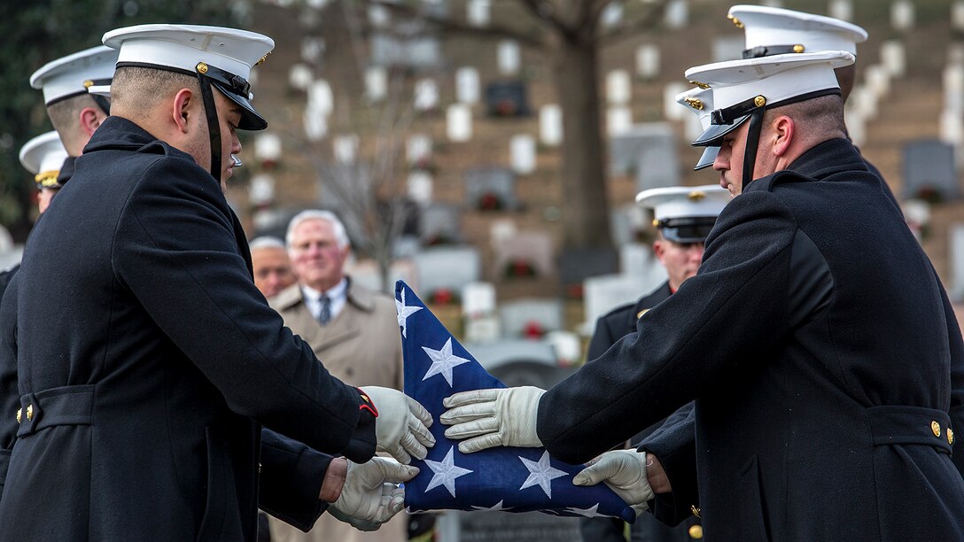Two Marines fold a flag between them into a triangle, with a backdrop of fellow Marines, onlookers and white headstones.