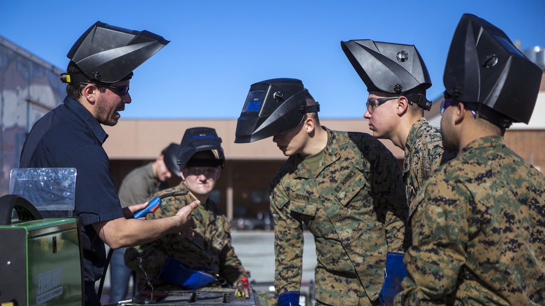Troops with pointy helmets open atop their heads, held at different angles, gather around welding equipment.