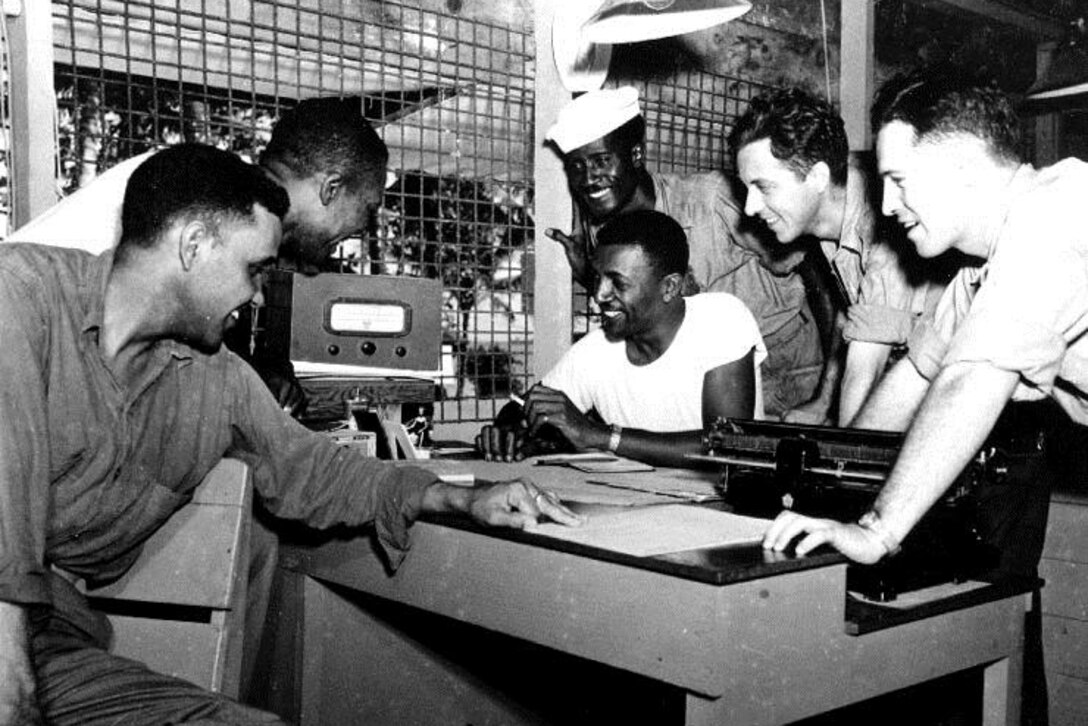 Men gather around a radio in a black and white photograph.