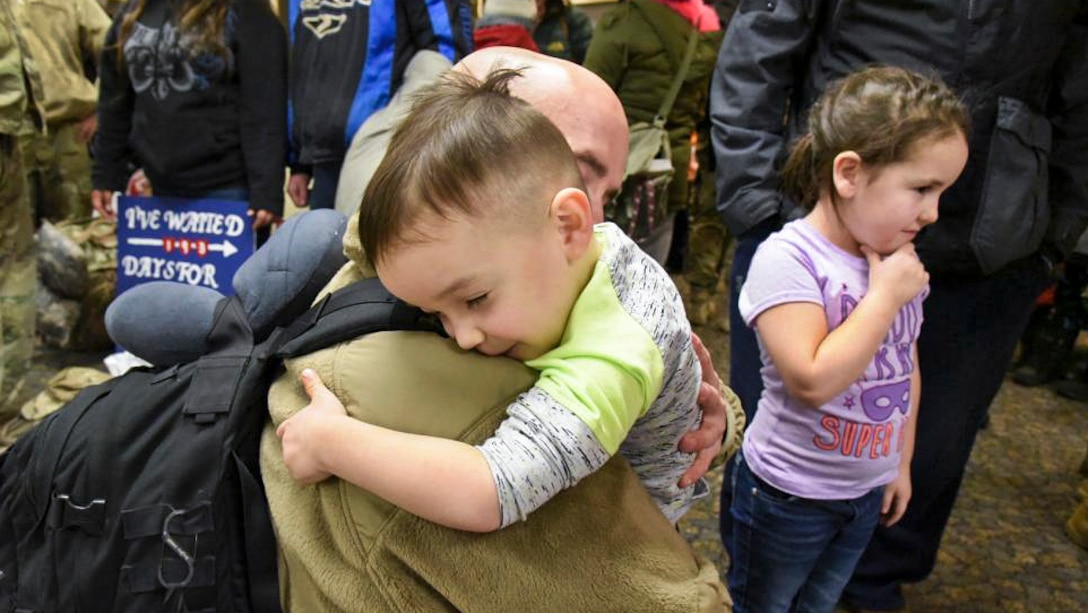 A boy, with closed eyes and a slight smile, tightly hugs his dad, who is kneeling to the boy's height and returning the embrace.