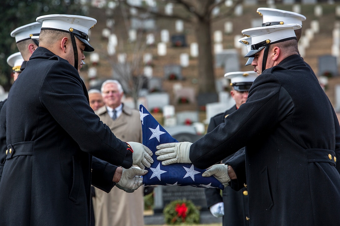 Two Marines fold a flag between them into a triangle, with a backdrop of fellow Marines, onlookers and white headstones.