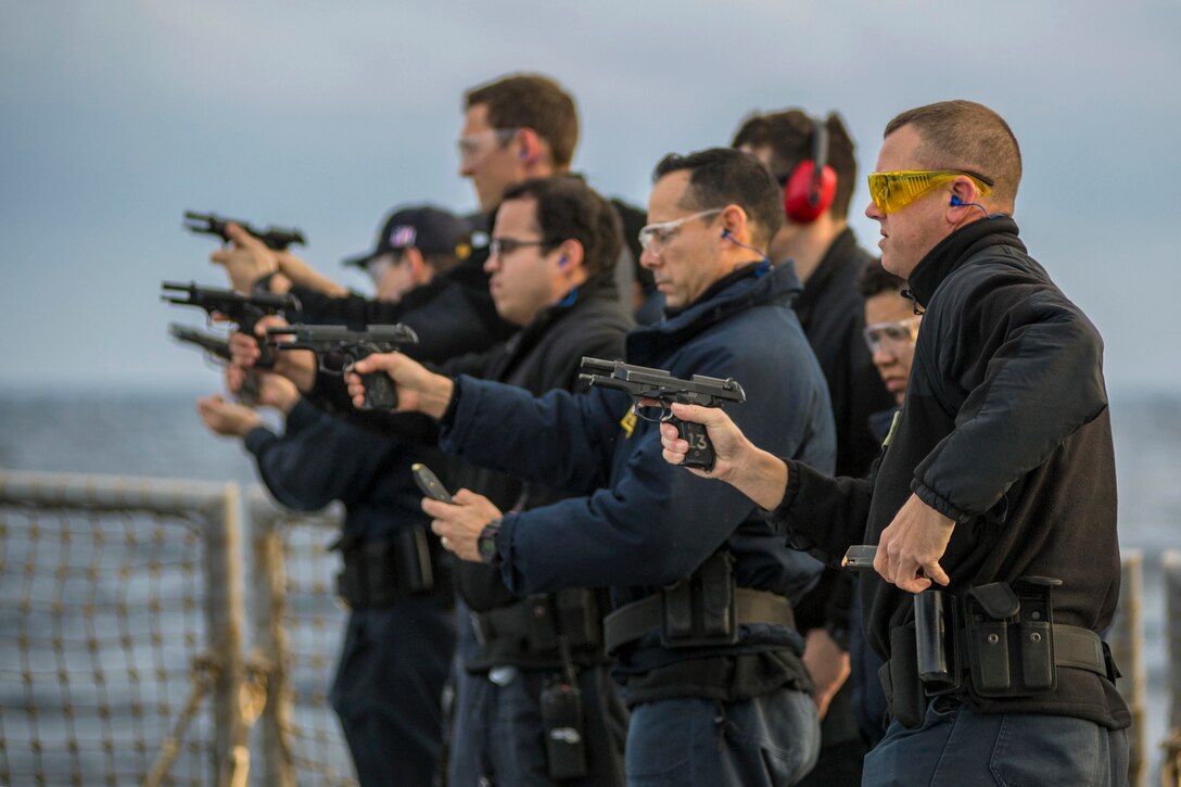 Sailors standing in a staggered row point pistols and hold ammunition.