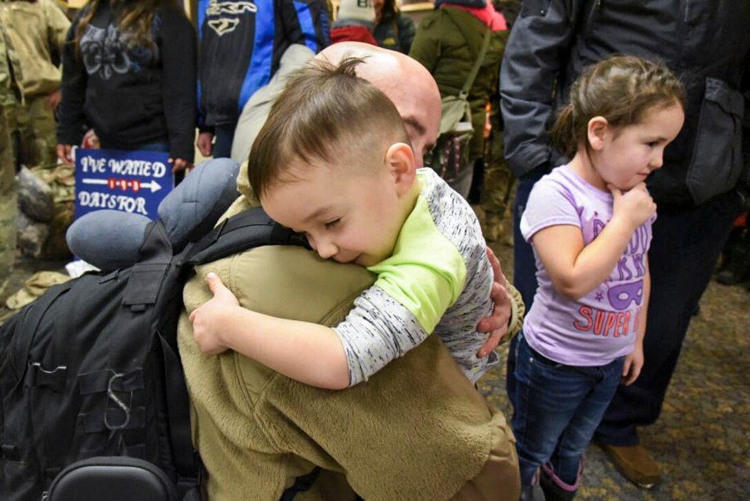 A boy, with closed eyes and a slight smile, tightly hugs his dad, who is kneeling to the boy's height and returning the embrace.