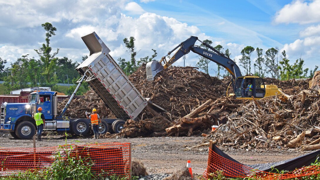 Workers operate a dump truck and tractor while moving a hill of vegetation.