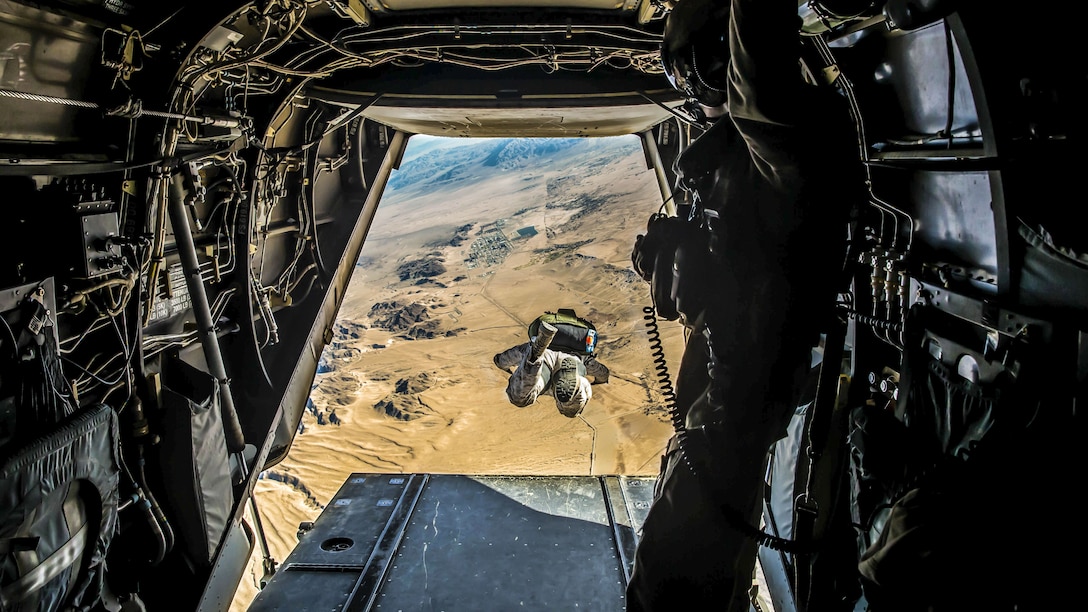 A Marine soars over desert terrain, visible from an opening in the aircraft from which he jumped.