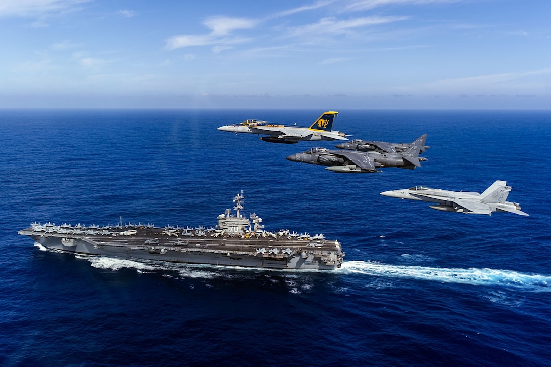 Four aircraft fly above an aircraft carrier traveling in blue sea.
