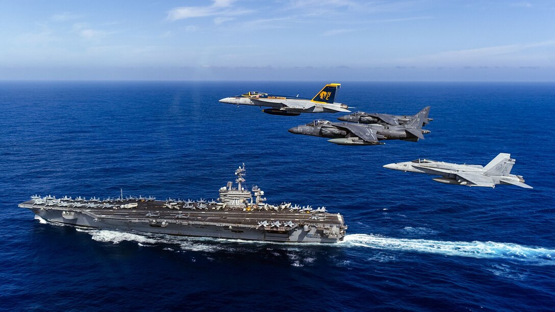 Four aircraft fly above an aircraft carrier traveling in blue sea.