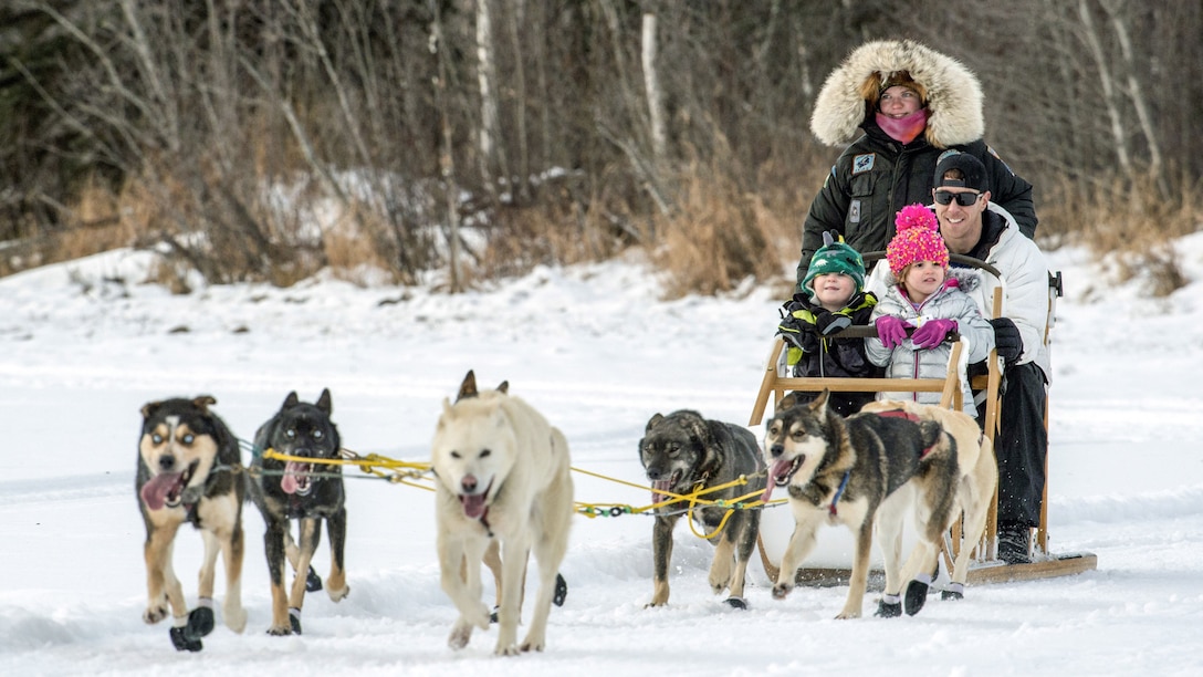 A man, two kids and a guide ride in a sled pulled by about six husky-type dogs.