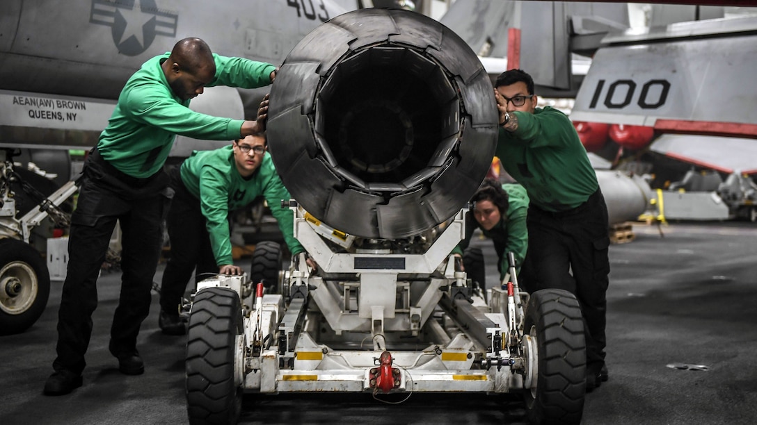 Four sailors in green shirts push an aircraft engine on a wheeled cart.