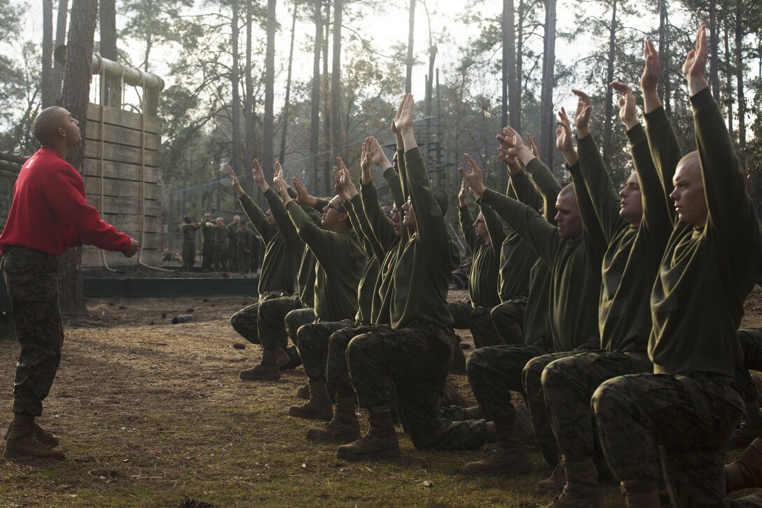 Marines kneel with hands stretched in the air with another Marine in front of them.