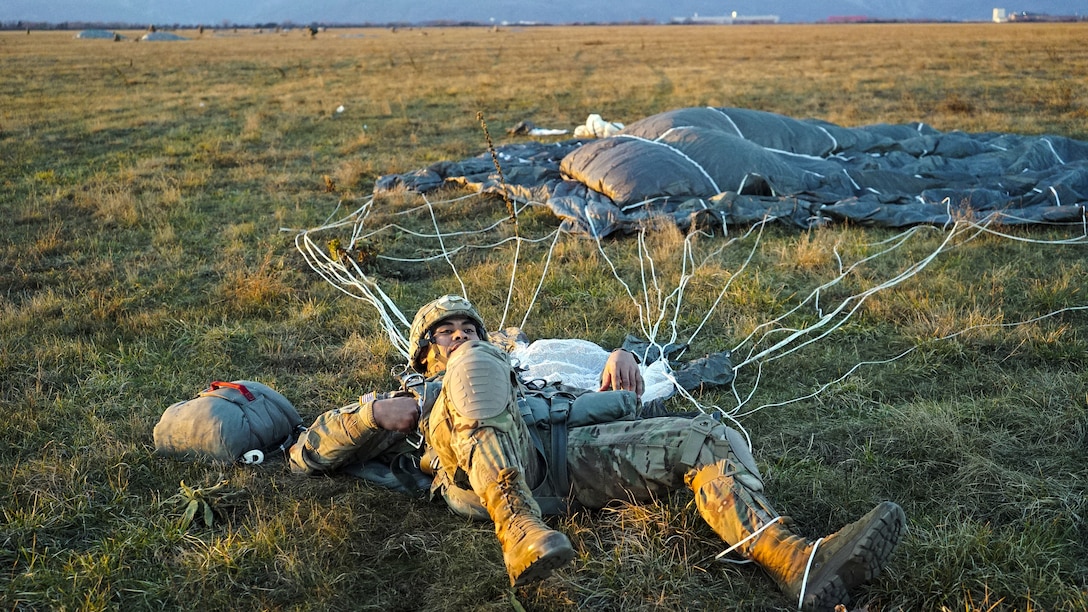 A soldier lies prostrate on the ground with his parachute spread out on the ground nearby.