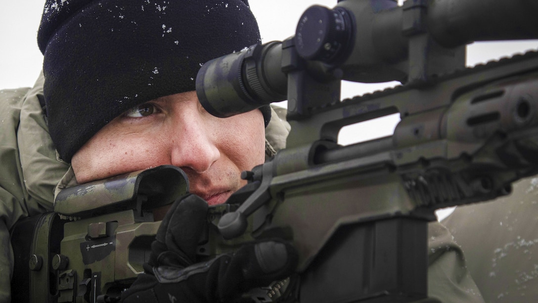 A sniper in a snow-spotted black hat looks through a rifle scope.