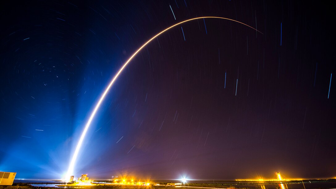 A curving beam of light illuminates a dark blue sky as a rocket launches.