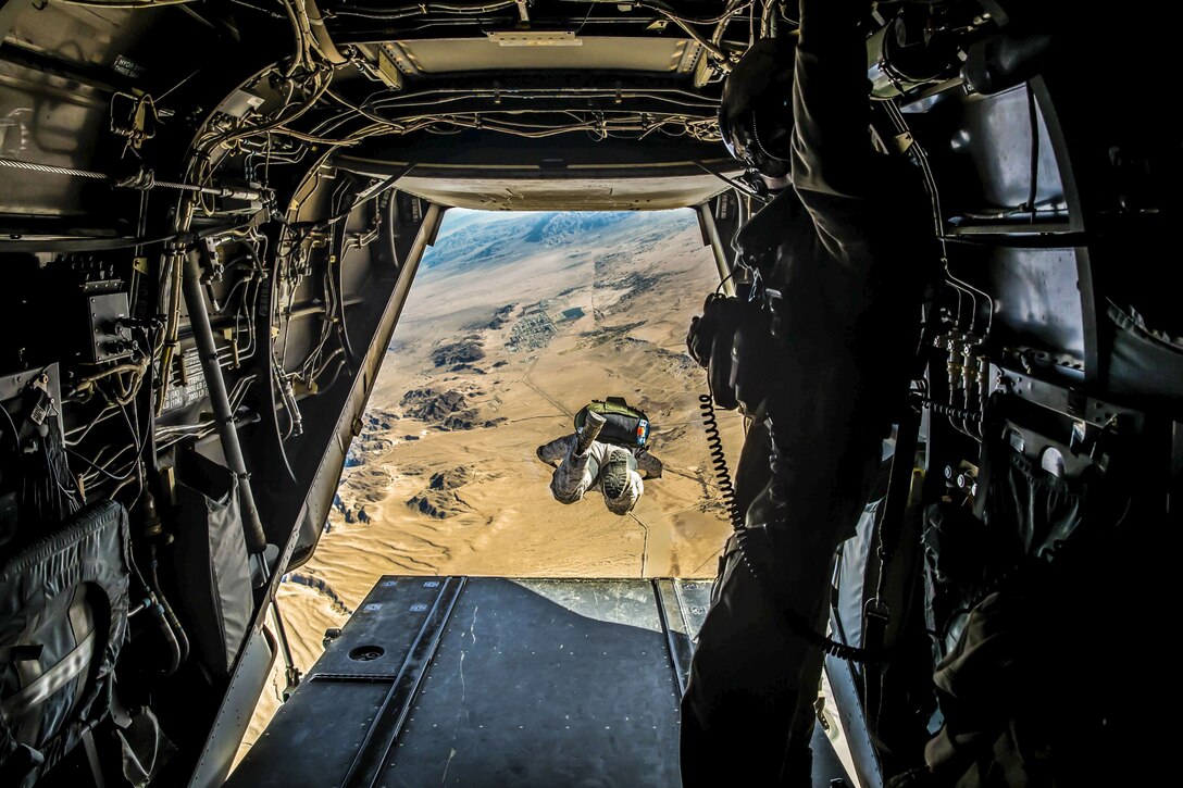 A Marine soars over desert terrain, visible from an opening in the aircraft from which he jumped.