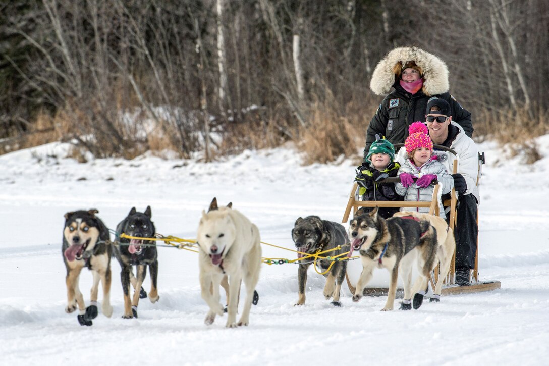 A man, two kids and a guide ride in a sled pulled by about six husky-type dogs.