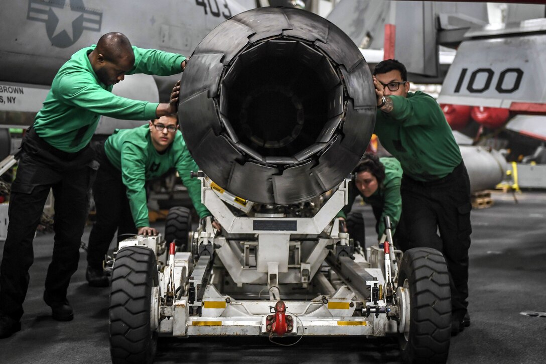 Four sailors in green shirts push an aircraft engine on a wheeled cart.