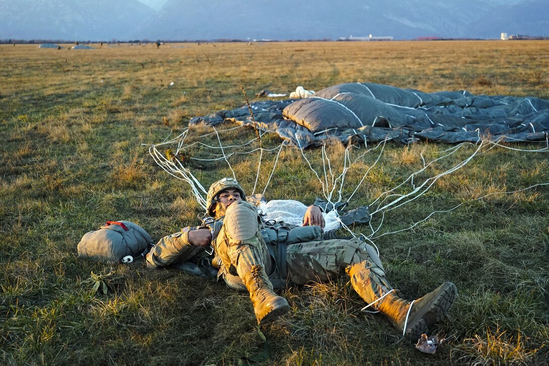 A soldier lies prostrate on the ground with his parachute spread out on the ground nearby.