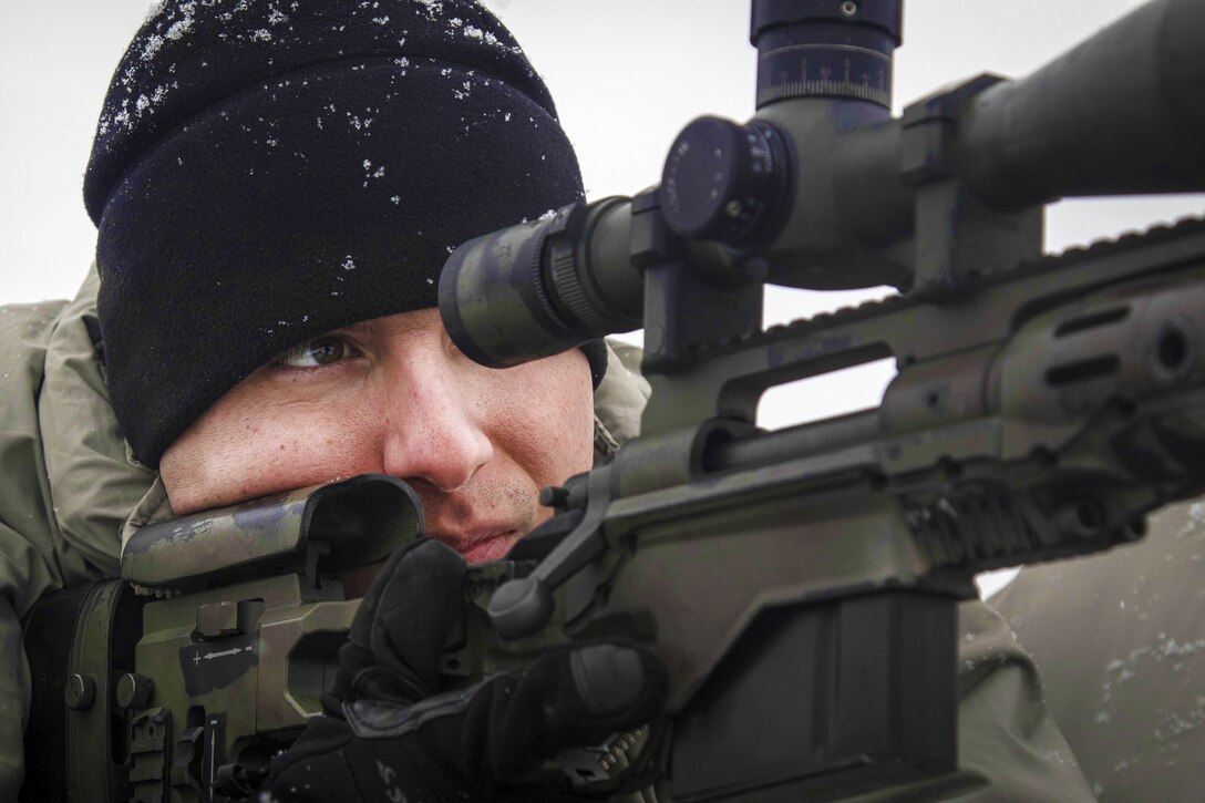 A sniper in a snow-spotted black hat looks through a rifle scope.