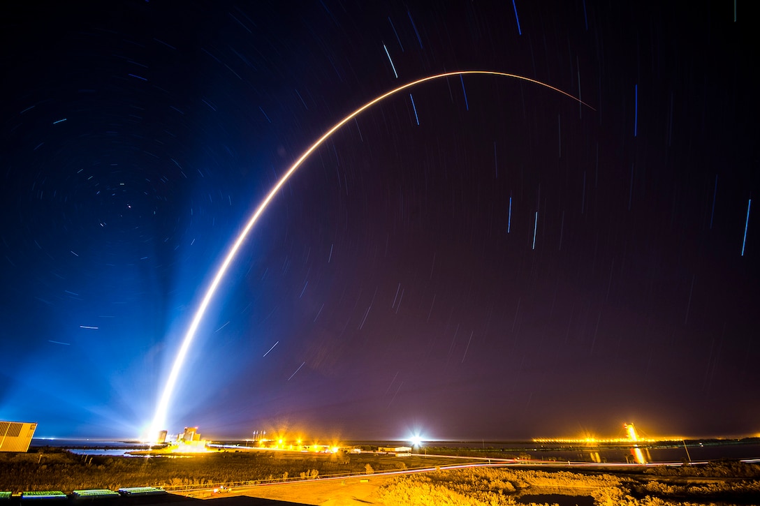 A curving beam of light illuminates a dark blue sky as a rocket launches.