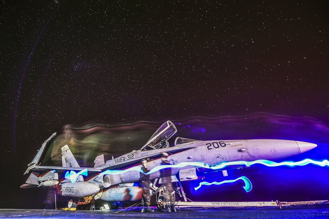 Wavy beams of colored light emanet from a jet on a flight deck at night.
