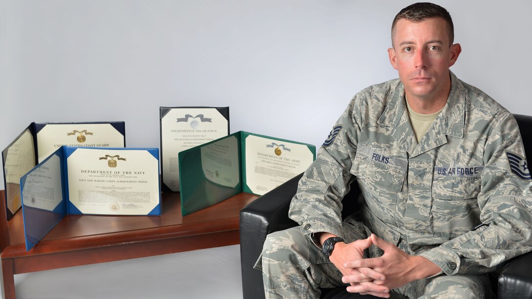 An airmen sitting next to four awards displayed on a table.