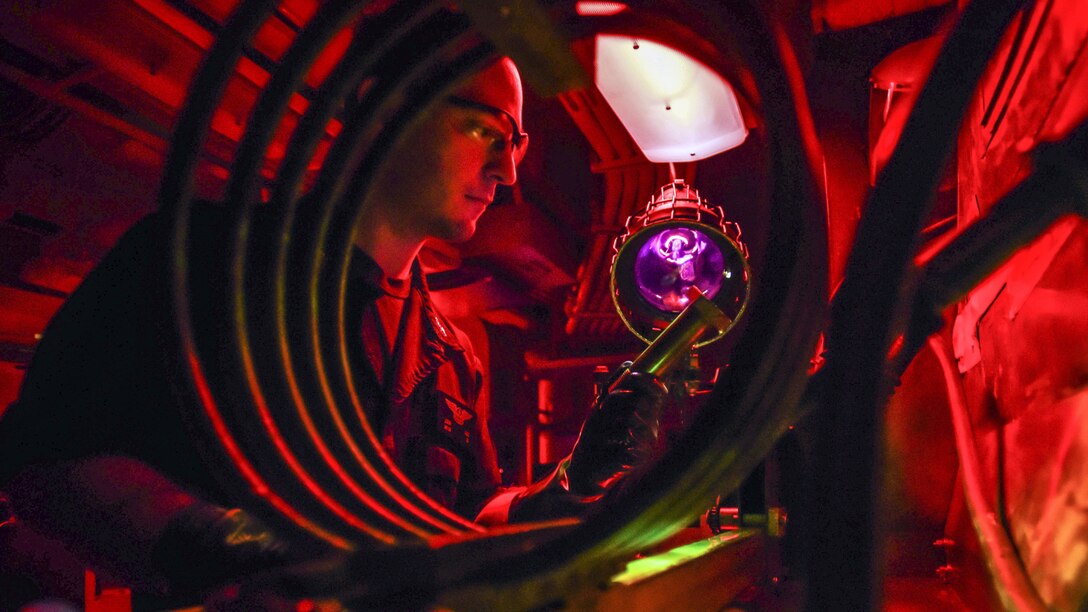 A sailor, illuminated in red and framed by a spiral structure, uses a light to look at an aircraft part he's holding.